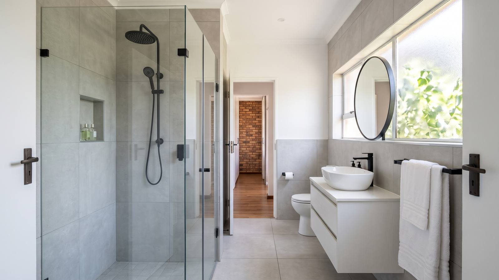 A newly renovated bathroom with a frameless glass walk-in shower, large grey porcelain wall tiles, a floating vanity and a window letting in daylight.
